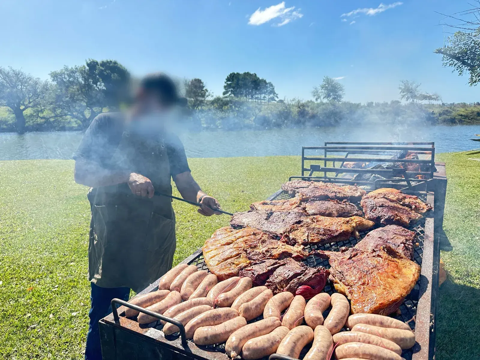 El asador cocinando bifes y chorizos en una parrilla grande, junto a una laguna